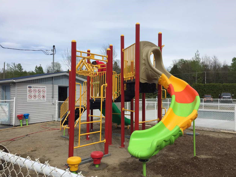 This community center outdoor playground features a KidsVenture Classic Series playset. It includes two elevated platforms, each with a vibrant, colorful slide that adds excitement and visual appeal. A compact monkey bar offers active climbing fun, while bright yellow railings provide safety and a cheerful touch. Several stepping pods connect the play elements, encouraging balance, coordination, and exploration.