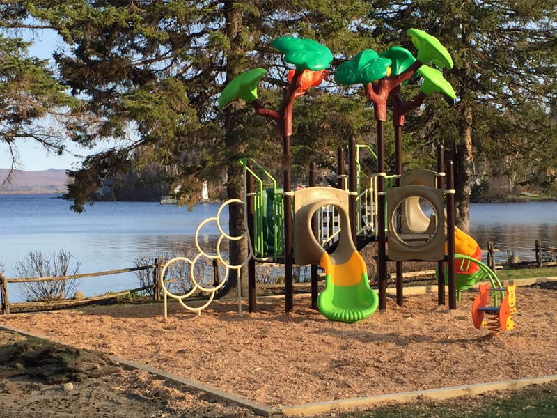 This nature tree outdoor playground sits right by a calm lake, surrounded by tall evergreens and fresh open air. The play structure blends right into the natural setting, with big, leafy tree canopies above and soft wood mulch underfoot. Kids can climb through circular rings, crawl through tunnels, and slide down smooth, colorful slides. The ground feels soft and springy, and the scent of wood adds to the outdoor experience. You can hear birds in the trees and water gently moving nearby—it’s a peaceful, playful place.