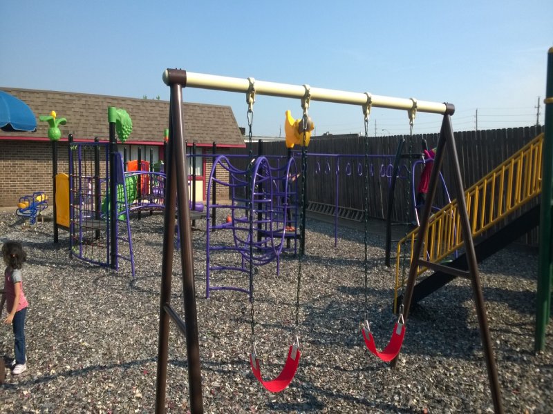 This custom-designed project was created for an elementary school outdoor playground. It features a variety of play elements suitable for children ages 2 to 12. A 5-foot-high standalone play structure with a bright yellow railing and a vibrant green spiral slide adds excitement for older kids. A classic monkey bar invites children ages 4 and up to test their courage and upper-body strength. Multiple net climbers challenge children of all ages to develop balance and coordination. The lower deck, equipped with two small purple parallel slides, creates a welcoming space for younger children to play together and build social skills. The inclusive design also ensures children with disabilities can enjoy outdoor play in a safe and engaging environment. Nearby, a swing offers younger children the thrill of motion while helping them bond with caregivers. This photo highlights the exhilarating swing.