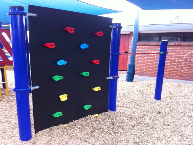 This climbing wall features a sleek, vertical black surface with vibrant handholds that add both function and fun. Two bright blue posts support the structure, tying it into the playground’s bold color scheme. It’s part of a custom-designed climbing circuit at an elementary school in Brisbane, Australia.
