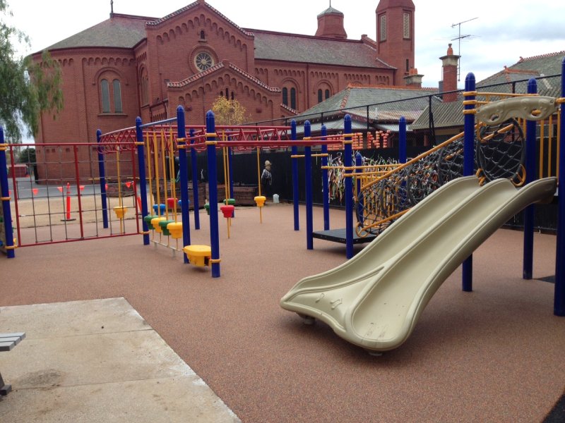 This custom-designed playground was created for an elementary school in Sydney, Australia, with a perfect balance of fun for all age groups. It features a variety of upper body challenges like monkey bars, a climbing wall, and several net climbers. We also included one of our signature KidsVenture play structures. Bright blue posts paired with bold red frames and rings give the space a vibrant, energetic feel. The main play structure extends the blue theme with a matching roof, yellow railings, and two slides — one open and one fully enclosed. The playground is built to engage kids from ages 2 to 12. Younger children can enjoy climbing and crawling on the play structure, while older kids can test their strength and coordination on the monkey bars and climbing wall. This photo captures the fun on this double slide.