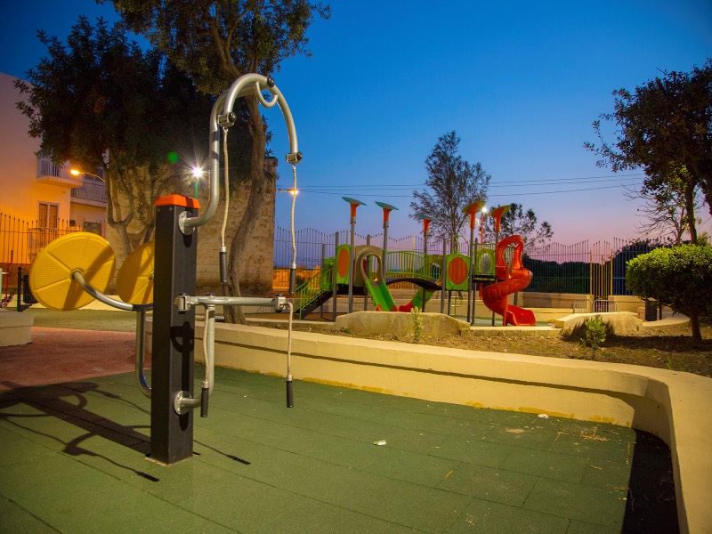 At the center of the design is a colorful, multi-functional playset featuring slides, climbing structures, and interactive panels. Surrounding the main structure is a collection of spring rockers in various shapes, offering playful motion-based activities for younger children. The entire area is safely enclosed and surfaced with impact-absorbing material to ensure both security and comfort for families. What sets this Malta project apart is its investment in outdoor fitness equipment. Strategically placed throughout the open space, the equipment includes hand bikes, elliptical walkers, leg presses, and upper-body stations.