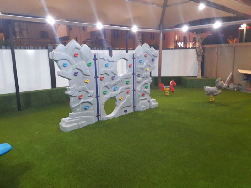 Colorful outdoor playground playset at a five-star hotel in Riyadh, featuring slides, climbers, steeping forms, net climbing, and artificial turf under a well-lit canopy. This photo highlights a custom designed artificial climbing wall.