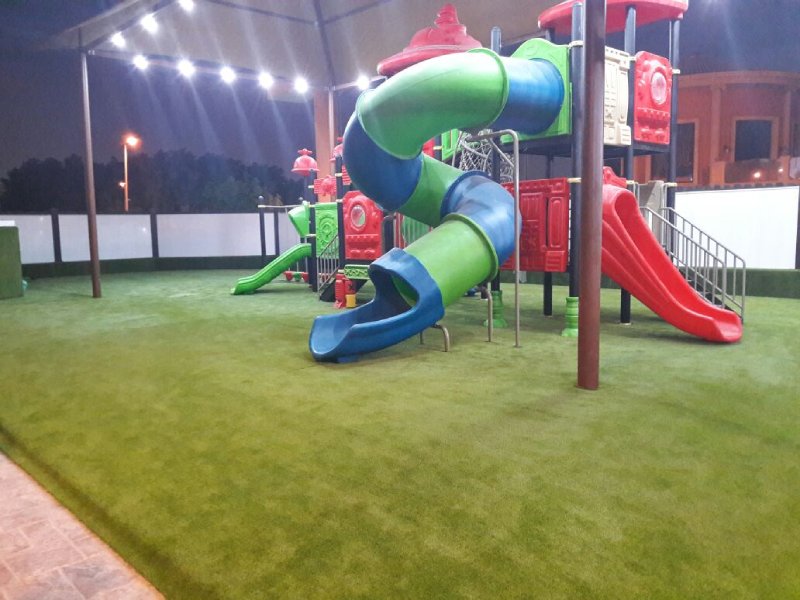 Colorful outdoor playground playset at a five-star hotel in Riyadh, featuring slides, climbers, steeping forms, net climbing, and artificial turf under a well-lit canopy. This photo captures the green and blue mixed enclosed spiral slide and a straight open red slide.