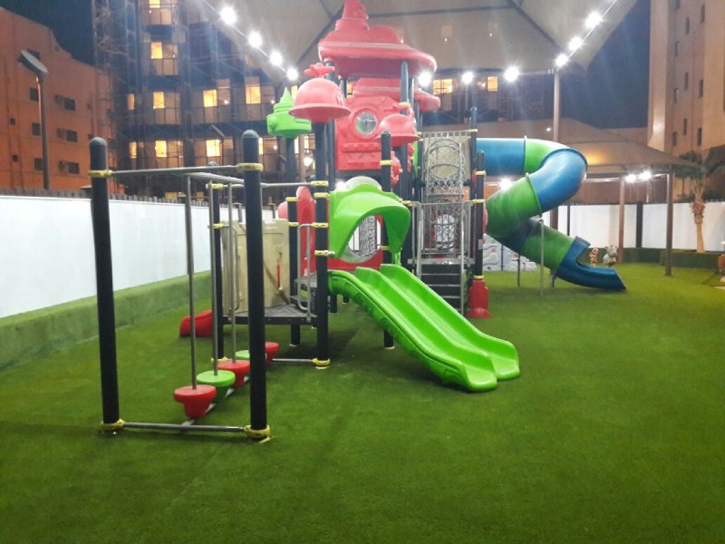 Colorful outdoor playground playset at a five-star hotel in Riyadh, featuring slides, climbers, steeping forms, net climbing, and artificial turf under a well-lit canopy. This photo captures the colorful stepping forms and bright green double slides.